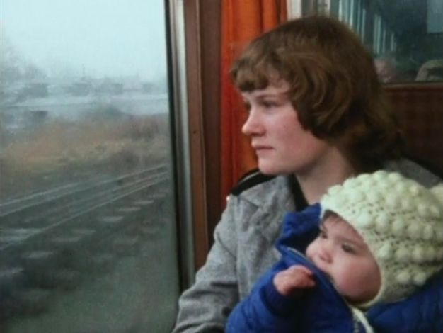 Video still. A pale-skinned woman and baby look out a train window in this grainy image. The woman’s brown hair and bangs frames her face. She has an upturned nose, and her closed lips turn slightly down. A fringe of dark hair peeks from under the knit hat the baby wears. Train tracks zoom by under a grey sky out the window.
