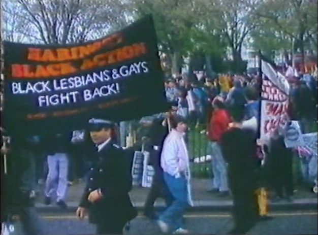 Video still. People gather, some walking, around a black banner with red and white text. Red text in all caps at the top reads “Haringey Black Action”. Below, white text reads, “Black Lesbians & Gays Fight Back!” A police officer stands between us and the crowd, which spill into a park or grassy area across the street from us.