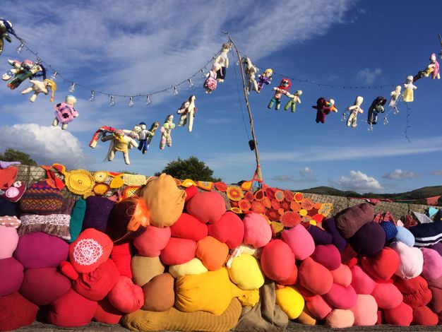 Installation photograph. Against a blue sky, sunlight pours onto two strings of unlit string lights and the two dozen sewn fabric votive dolls hung below them. Below is what appears to be a wall like that made with sandbags but with brightly coloured red, pink, and yellow pillows.