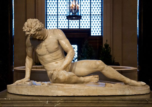 Sculpture. This freestanding statue shows a nude man sitting with his right leg tucked under him, the other extended. He leans on his right hand, and rests his other on his right thigh. He looks straight down so we see his furrowed brow, nose, and a hint of his moustache. A rings that opens at the front is around his neck, and blood dribbles out of a wound over his right ribs.