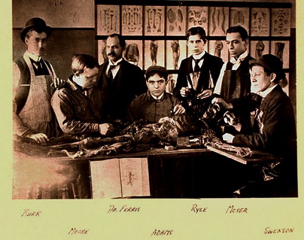Black and white photograph. Seven men gather around a skeletal cadaver. The arm we can see is stretched along a board. The wall behind the group is hung with diagrams and illustrations. The individuals are labelled in handwriting under the photograph: Burr, Moore, Dr. Ferris, Adams, Ryle, Moser, and Swenson.