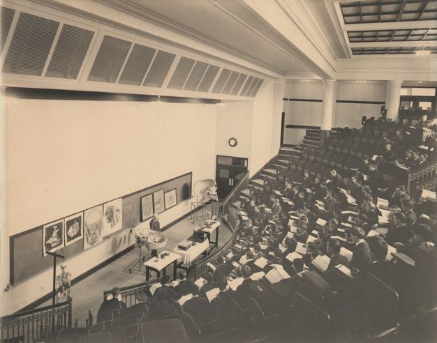 Black and white photograph. From a back row off to one side, we look down at about twelve rows of seats curving around a long blackboard hung with illustrations and diagrams in this lecture hall. A man stands between the blackboard and tables lined with specimens and materials. Students fill the seats down the centre of the hall.