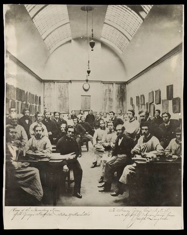 Black and white photograph. About forty men pose in a hall-like dissecting room. The ceiling is rounded, like a tunnel, and set with skylights. A pair of feet hang off the end of the table closest to us to our left and a skull sits at the front edge of the table to our right.