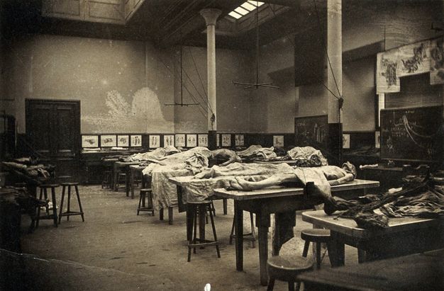 Black and white photograph. About a dozen cadavers are laid on high, long tables. Some are covered or partially covered with a sheet.