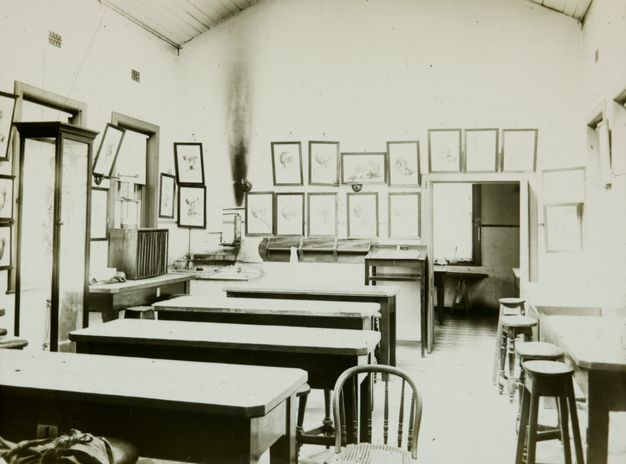 Black and white photograph. Six tall, long tables are laid out in a room hung with framed illustrations of dissections.