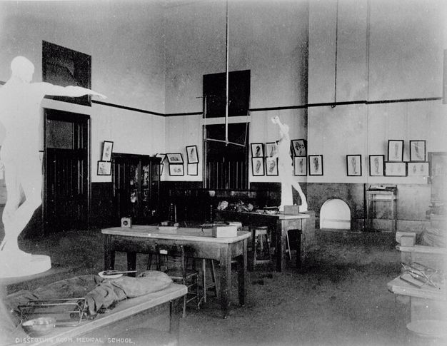 Black and white photograph. Two white statues of men in a classical style loom over long tables set with equipment in this empty, hall-like room.