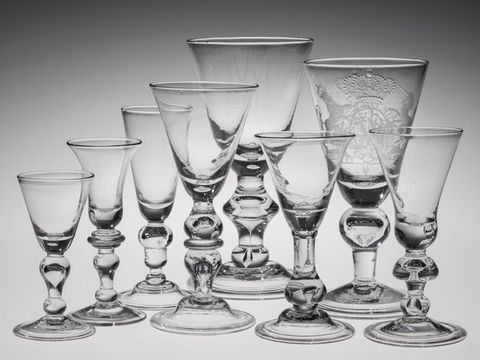 Glass. Eight clear glass drinking vessels are shaped like upside-down triangles or bells. Each has a knob on a thick stem over a flaring foot. Most of the knobs are right under the bowl of the glass but one has a knob closer to the foot.