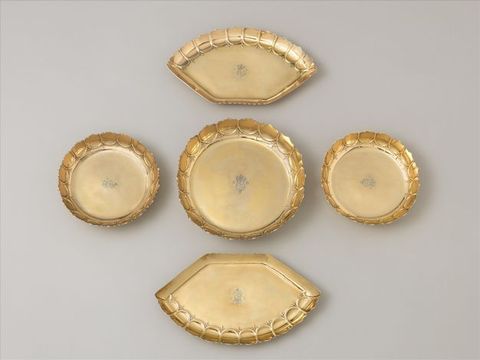 Metalwork. Five gold dishes with fluted, scalloped edges are stamped with a coat of arms and the letters “GR”. Three are round, and one is larger than the other two. The final two are flat on one side and curve in a segment of an arch on the opposite side.