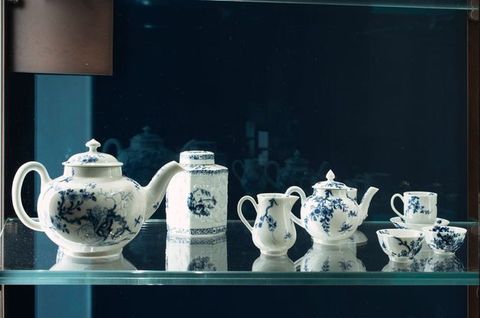 Installation. Two teapots, a jug, three cups, and a canister painted with blue designs against white porcelain are in a case. They reflect in the glass on the far side.