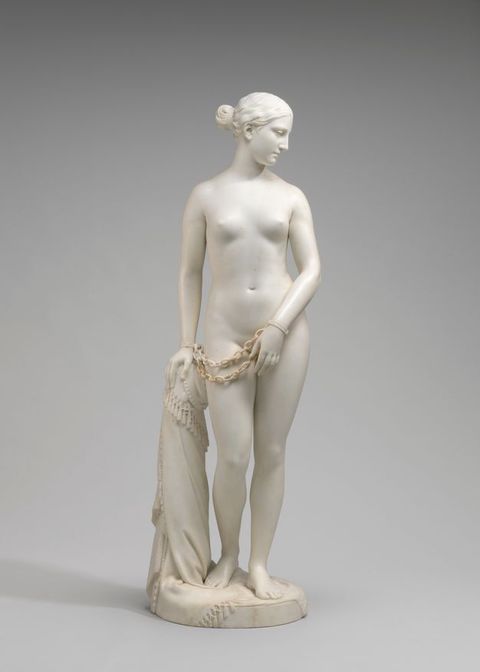 Sculpture. This free-standing statue of a nude woman standing with one hand on a hip-high post, her body facing us as she looks down and to our right in this view. Chains hang from shackles around her wrists.