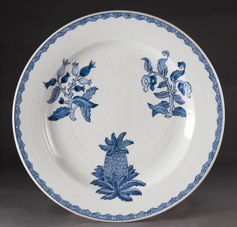 Porcelain. A white plate is painted with two specimens of flowers and one pineapple. The edge has a narrow, swirling geometric pattern.