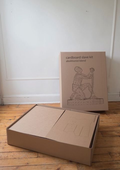 A box holding sheets of cardboard sits on a wood floor in front of the lid, which is propped on the wall behind it. The lid has the title printed above a line drawing of the sculpture the buyer can build with the kit.