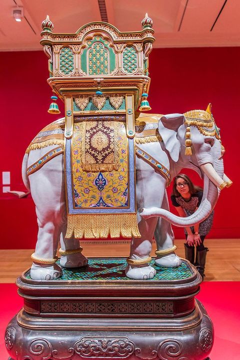 Sculpture. An elephant is draped in gold cloth patterned with blue and red below a balcony-like structure called a howdah. A woman peering through the oval space left by the elephant’s trunk gives a sense of scale. The complete earthenware sculpture is about half again as tall as the woman would be.