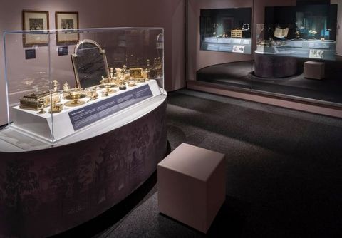 Installation. A long, oval-shaped platform holds a case filled with gold-coloured glass vessels, boxes, and a mirror.