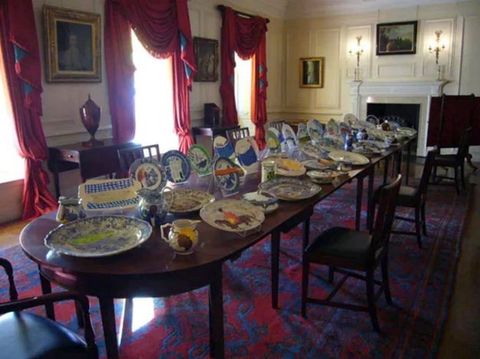 Installation. Plates and dishes are set and displayed on a long dining room table. Pale yellow panelling covers the walls and heavy scarlet-red drapes hang from the windows. Details on the plates are difficult to make out but many are colourful and are different sizes.