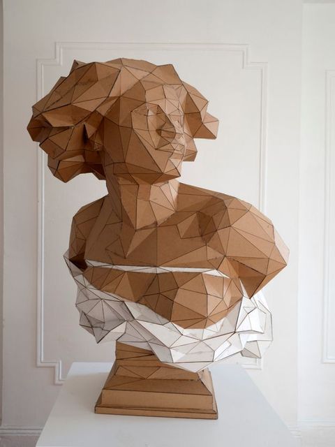 Sculpture. The head, shoulders, and chest of a Black woman are made with triangular and trapezoidal pieces of cardboard. In this view, her shoulders square towards us and she looks off to our right. Her skin, hair, and the base are the brown of the cardboard. White pieces make a rope across her chest and drapery below.