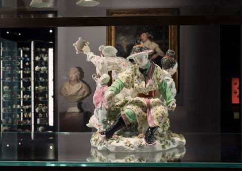 Porcelain. A grouping of at least three adults and one child are in a ring of activity and movement. One person holds a tambourine out, and the child holds up a small object as a man, closest to us, grips the child’s other hand. The porcelain is glazed with pastel yellow, spring green, rose pink, red, and black.