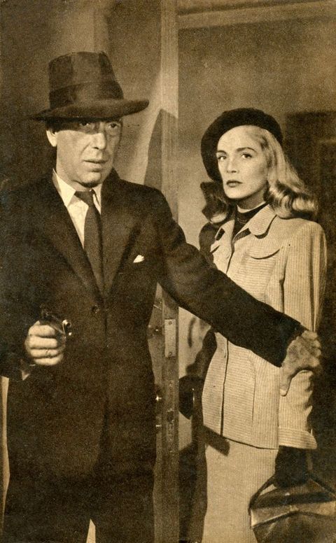 Film still. Black and white image of a pale-skinned man and woman standing near an open door. The man wears a suit and fedora, and holds up revolver in one hand. His other hand reaches across the woman’s body and clutches her far arm. She has long, shiny blond hair under a black cap, and wears a skirt suit and black gloves.