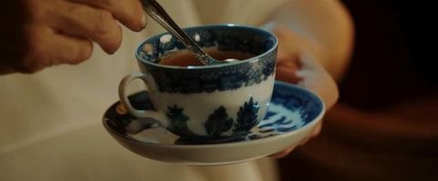 Video still. Close-up view of a blue and white porcelain teacup. A pale-skinned hand holds a silver spoon in the brown tea.