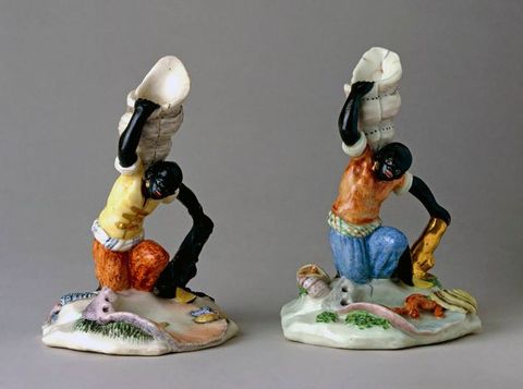 Porcelain. Two candlestick holders are in the shape of people with black-coloured skin kneeling and holding up seashells. The eyes are painted white, the mouths red, and each has a gold earring in the ear we can see.