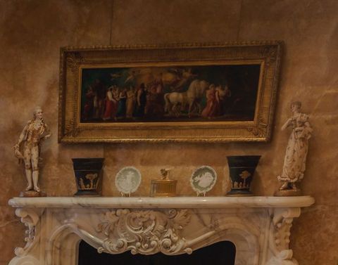 A long, gold-framed painting hangs over an ornately carved marble fireplace. Porcelains and ceramics are lined up on the mantle.