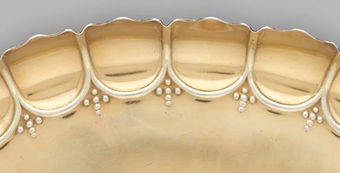 Metalwork. Detail view of a scalloped, fluted edge of a gold-coloured dish. Each scallop is ridged along its inner edge, and an ornament made up of six dots creating a cross is tucked into the space where the curves meet.