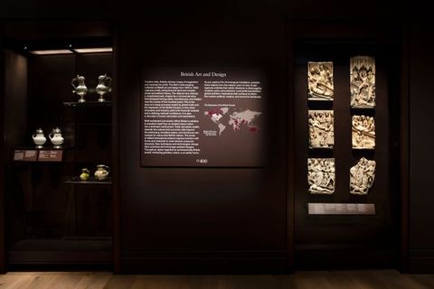 Installation. A wall text with white lettering on a dark background is headed “British Art and Design”. The text cannot be made out from where we are, but there is a map with markings in plum purple and white. A display case with porcelain, ceramics, and metalwork is to our left, and six relief panels to our right.