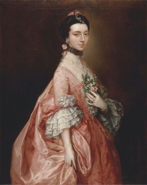 Painting. Shown from the thighs up, a pale-skinned woman wearing a carnation pink dress with cascading lace cuffs at the elbows stands in front of a dark background. Her body faces our right, her far hand resting on a spray of pink and white flowers and green leaves at her chest.