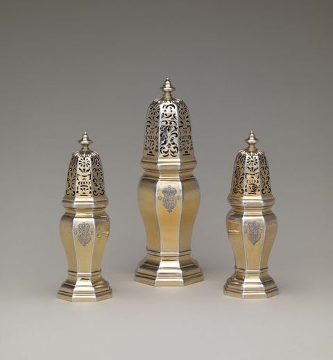 Metalwork. Three different sized casters would have been used like salt and pepper shakers but for sugar, dry mustard, and pepper. Each has an elongated top pierced with scrolls and floral designs. The bodies are six-sided, and each is incised with a coat of arms and the letters “GR”. Some of the gold surface has worn away to show the silver beneath.