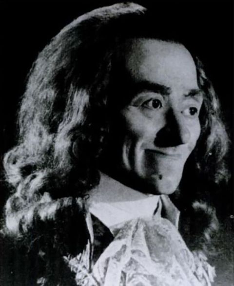 Black and white photograph. A man with thin lips pressed together in a smile looks off to our right. He has thick, dark brows, and his long hair hangs loose. He wears a high-collared white shirt and a frilly cravat.