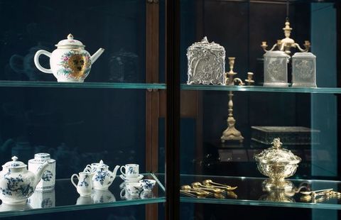 Installation. Four glass shelves in a cabinet hold porcelain, silver, and gold vessels, containers, and utensils for storing and making tea. The objects reflect in the glass on the back side and in the glass of the shelves, creating double and sometimes triple vision.