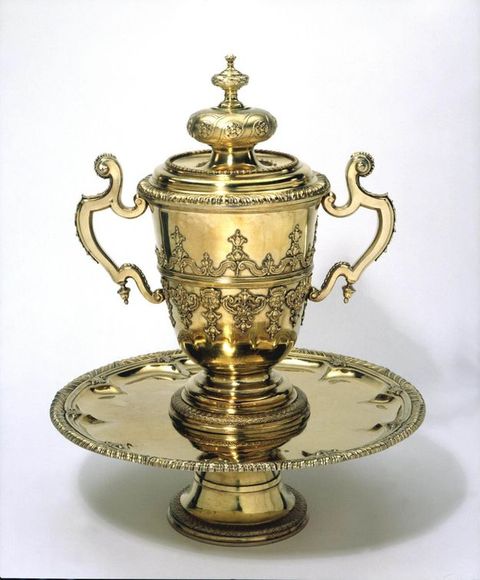 Metalwork. A lidded cup sits on a circular, footed tray. All three pieces are coloured gold. The cup has two scroll handles, and the body of the cup is decorated with strapwork and ornamental designs in relief. The lid has a bulbous and then smaller knob beneath a delicate finial.
