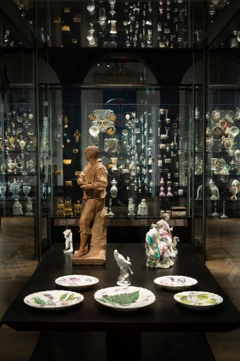 Installation. Statuettes of people and a bird are in a case with five plates. The wall opposite us is filled with porcelains and metalwork. Repetitions and reflections in the glass create a jewellery box-like effect.