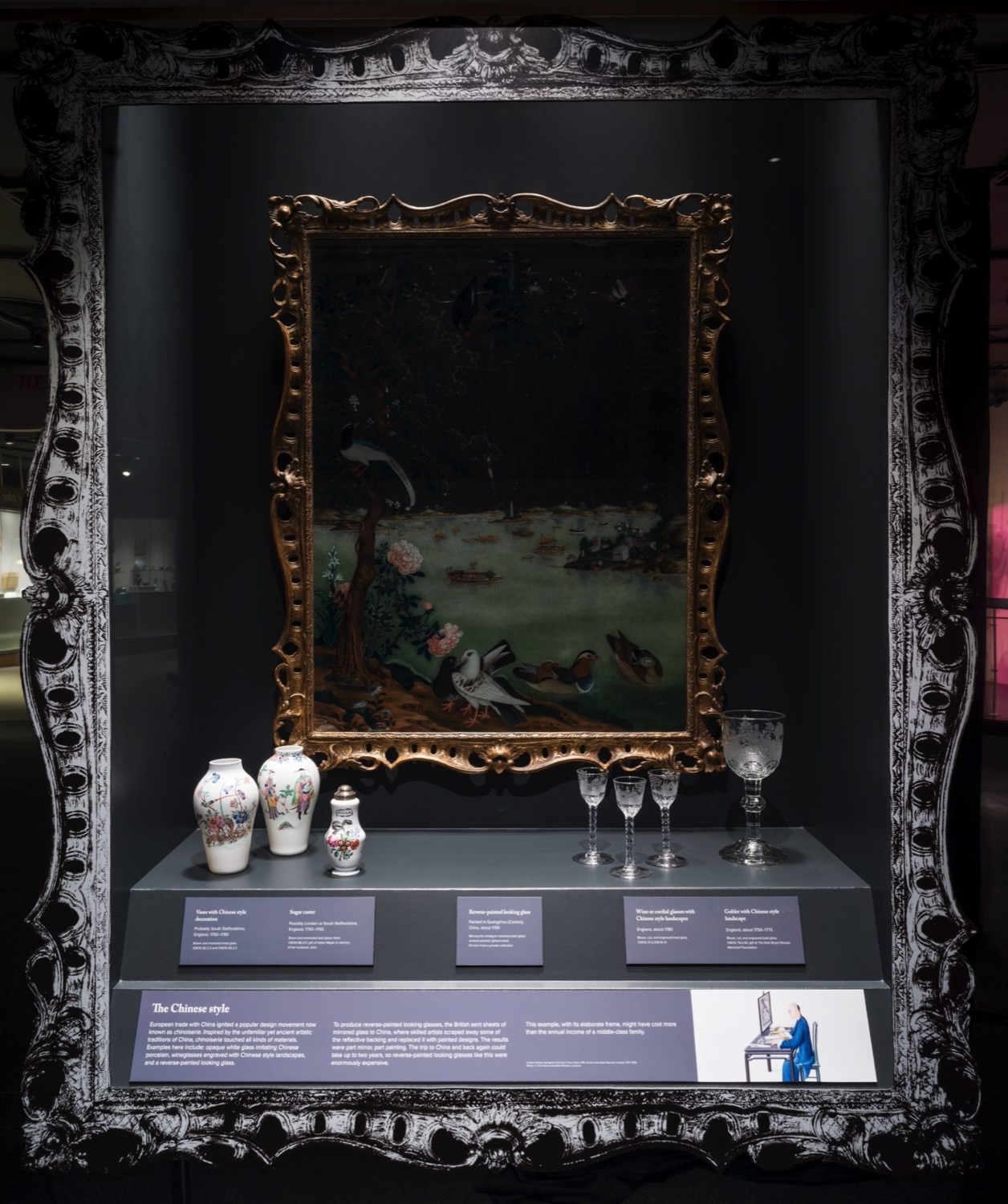 Installation. Porcelain vases, glasses, and a painting in a gold frame are in a case, which is, itself, framed with a duplicate of the gold frame, but in silvery tones.