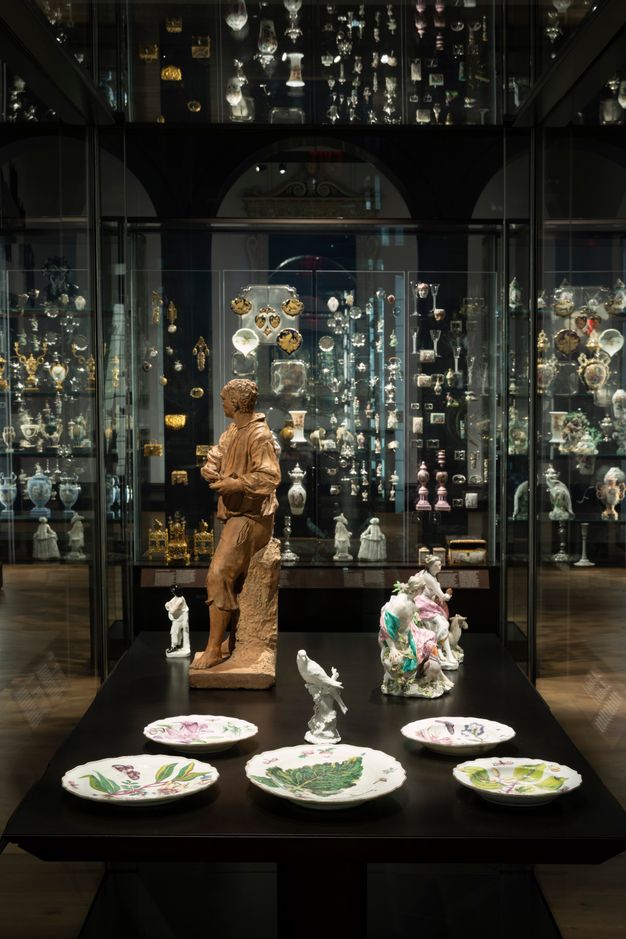 Installation. Statuettes of people and a bird are in a case with five plates. The wall opposite us is filled with porcelains and metalwork. Repetitions and reflections in the glass create a jewellery box-like effect.