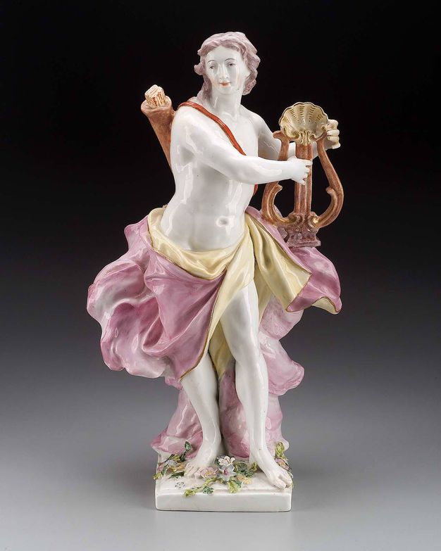 Porcelain. A man turns his shoulders to our right to hold and touch a lute, as he turns his head back to look off to our left. His skin is the white of the porcelain, heightened with pink on his cheeks and lips. A quiver of arrows is slung across his bare chest, and yellow and pink drapery flutters around his hips.
