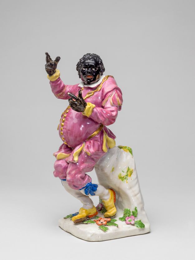Porcelain. A man with black-coloured skin wears a bubble gum-pink suit lined with yellow and with a row of gold buttons down his bulging belly. He leans back on a projecting ledge, his canary-yellow shoes tucked back under the ledge. Both hands are held up. The inside of his mouth is painted red, and the whites of his eyes stand out in the dark face.