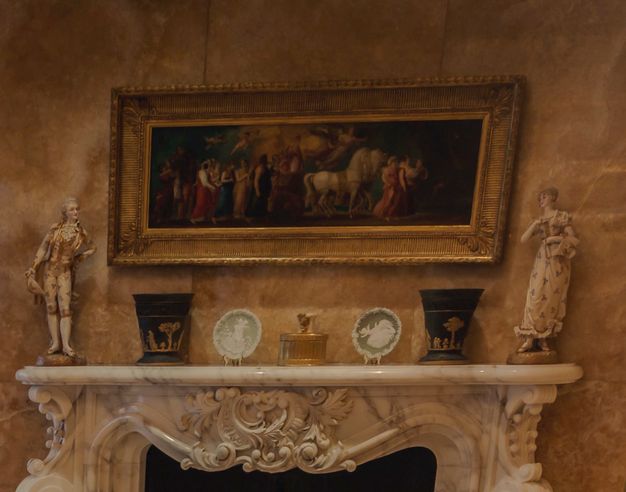 A long, gold-framed painting hangs over an ornately carved marble fireplace. Porcelains and ceramics are lined up on the mantle.