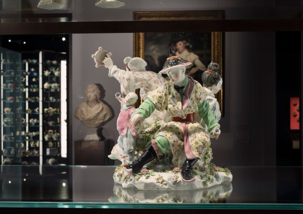 Porcelain. A grouping of at least three adults and one child are in a ring of activity and movement. One person holds a tambourine out, and the child holds up a small object as a man, closest to us, grips the child’s other hand. The porcelain is glazed with pastel yellow, spring green, rose pink, red, and black.