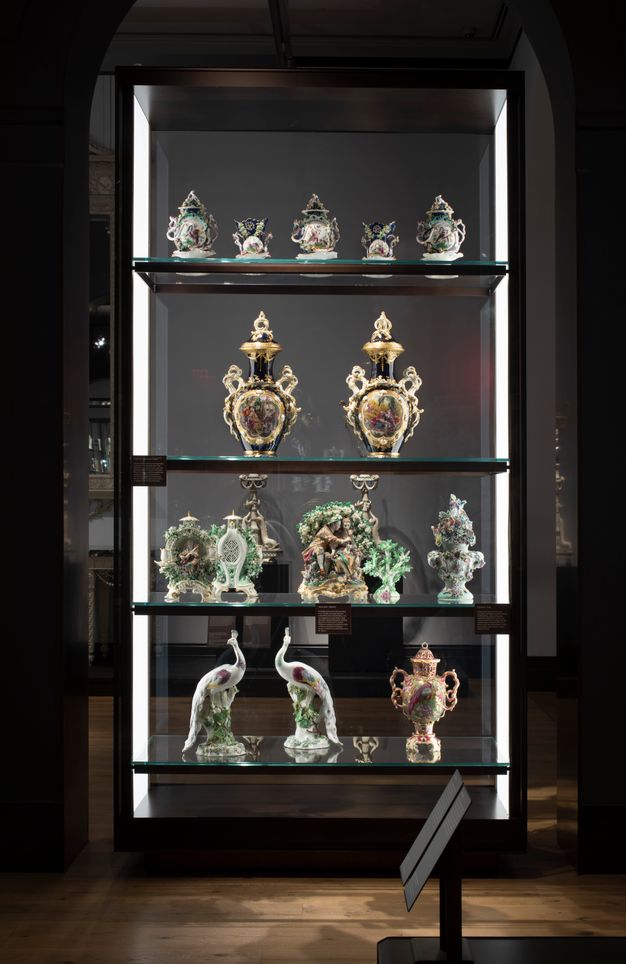 Installation. Four shelves are lined with glass on both long sides so we look through it. From the bottom up are a pair of birds on the bottom shelf and figural groups on the second shelf. Urns and vessels fill the rest of the space.