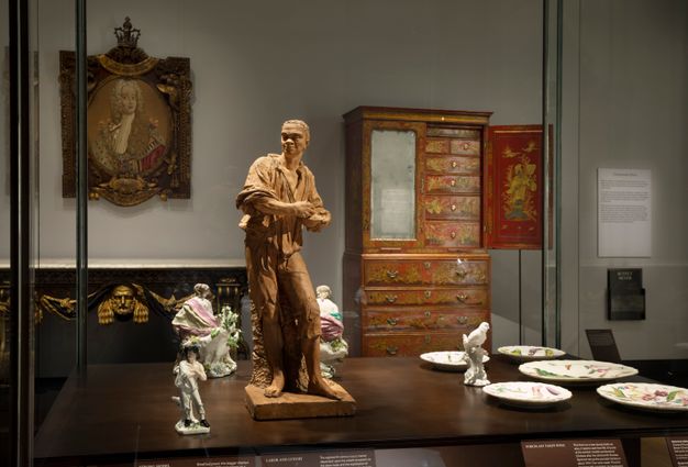 Installation. A case has four statuettes of people, one bird on a tree trunk, and five plates. A sideboard, chest of drawers, and a painting line the wall on the far side of the room.