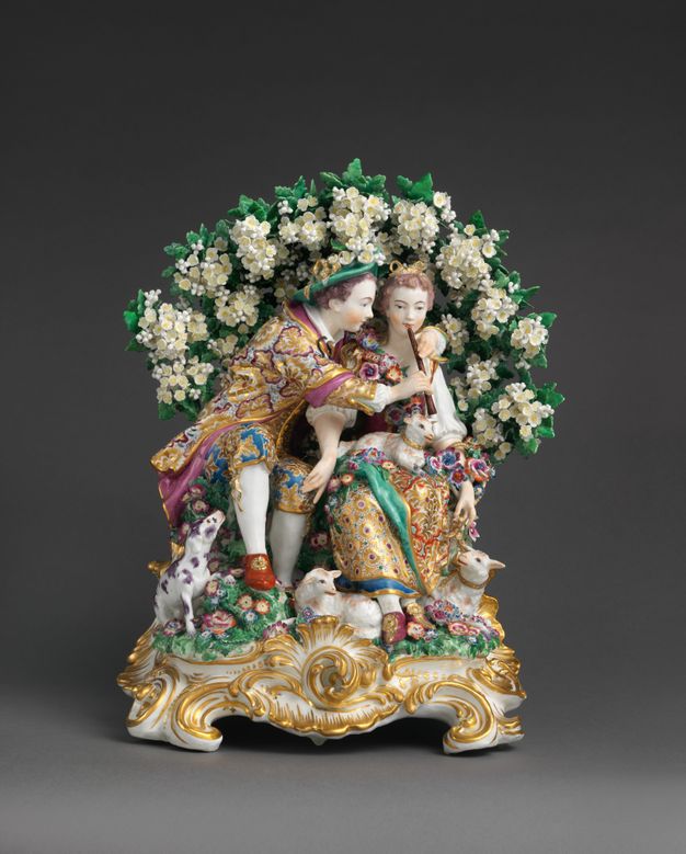 Porcelain. A young man and woman sit under an arch of white flowers. Both have pale skin tinged with peach on the cheeks and hands. The boy wraps both arms around the girl’s shoulders to hold a flute to her lips. She touches the front of one his knees with one hand.
