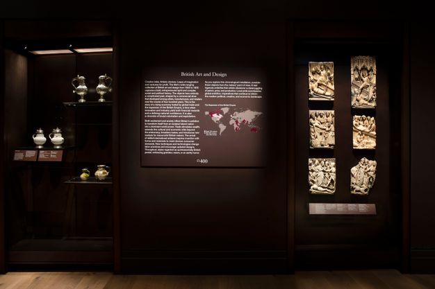 Installation. A wall text with white lettering on a dark background is headed “British Art and Design”. The text cannot be made out from where we are, but there is a map with markings in plum purple and white. A display case with porcelain, ceramics, and metalwork is to our left, and six relief panels to our right.