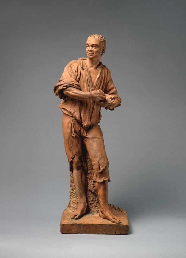 Sculpture. This freestanding terracotta statuette shows a Black man with tattered clothing and bare feet. He holds a bowl with muscular arms and large hands over one hip. His head turns back the other way and he smiles. He has short, curly hair, deepset eyes, and a wide nose.