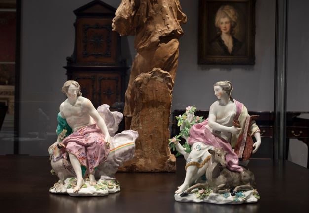 Porcelains. Two free-standing statuettes show a seated man and woman. Their skin is the white of the porcelain tinged faintly with peach, and they have ash-brown hair. The man is on a cloud streaked with a rainbow, and he rests his hand on a bird of prey. The woman holds a lute, and a stag lies at her feet.