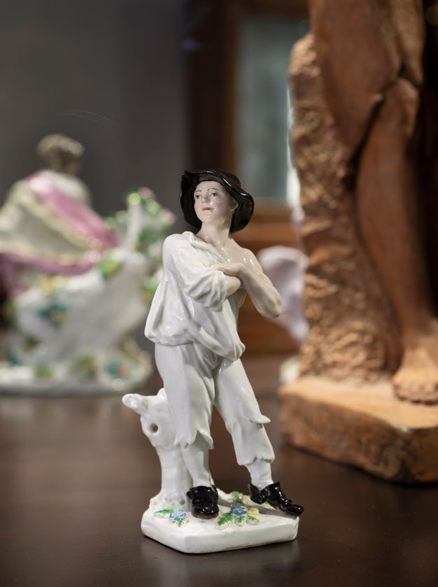 Porcelain. A figurine of a pale-skinned man stands with arms crossed, looking off to our left. He wears a wide-brimmed black hat, white, tattered clothes, and black shoes.