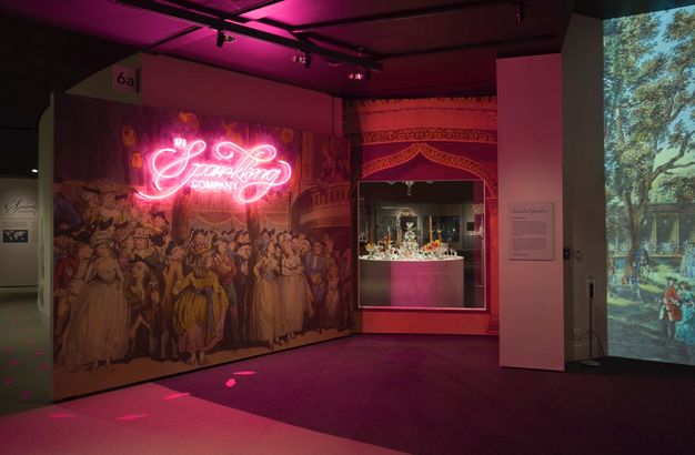 Installation. A floating wall is covered with an enlarged print showing women wearing long gowns and men wearing hats and suits. The title of the exhibition is in pink neon, illuminating the otherwise dim space. Pink-glowing footsteps seem to emerge from the print and turn the corner, leading us into the exhibition.