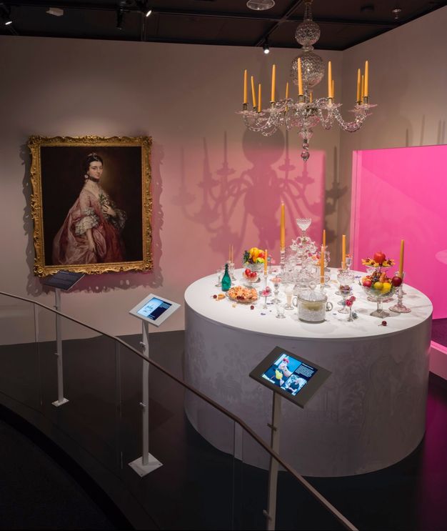 Installation. A round white platform holds glasses, candlesticks, bowls of fruit, and containers under a chandelier, all of which are glass (except the platform). A painting of a pale-skinned woman wearing carnation pink is framed in gold on the far wall. The space is dimly lit except for a fluorescent-pink glow from a case behind the platform.