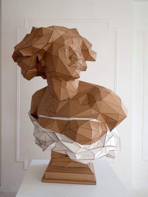Sculpture. The head, shoulders, and chest of a Black woman are made with triangular and trapezoidal pieces of cardboard. In this view, her shoulders square towards us and she looks off to our right. Her skin, hair, and the base are the brown of the cardboard. White pieces make a rope across her chest and drapery below.