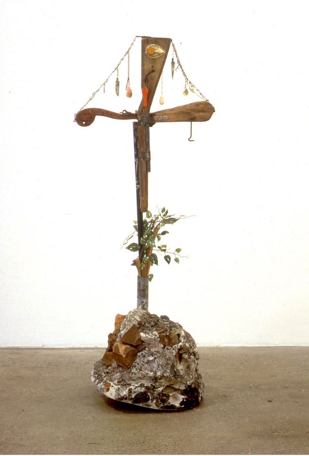 Sculpture. A rifle stands up from a concrete and brick base. Two pieces of wood make the rifle into a cross below the stock. Chains hanging from the top to the outer arms of the cross are hung with seashells.